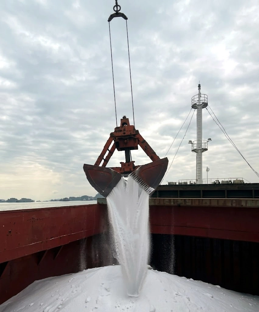Grab bucket pouring white bulk cargo into a vessel hold